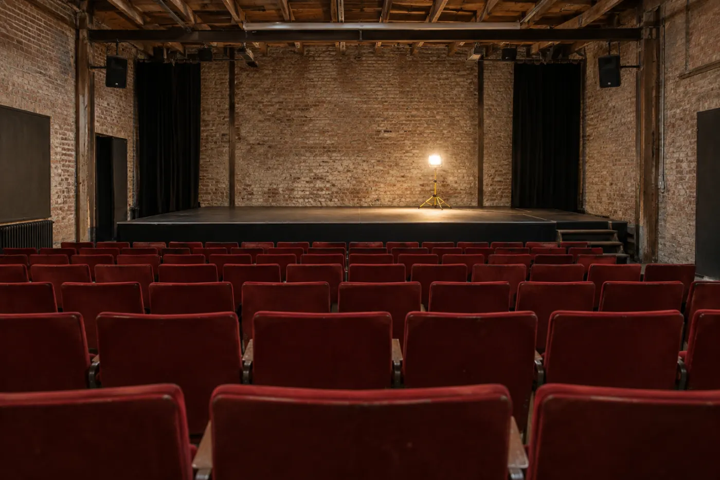 The 180-seat house from the back row: rows of red cinema seats, brick walls and timber beams, one work light on the bare stage