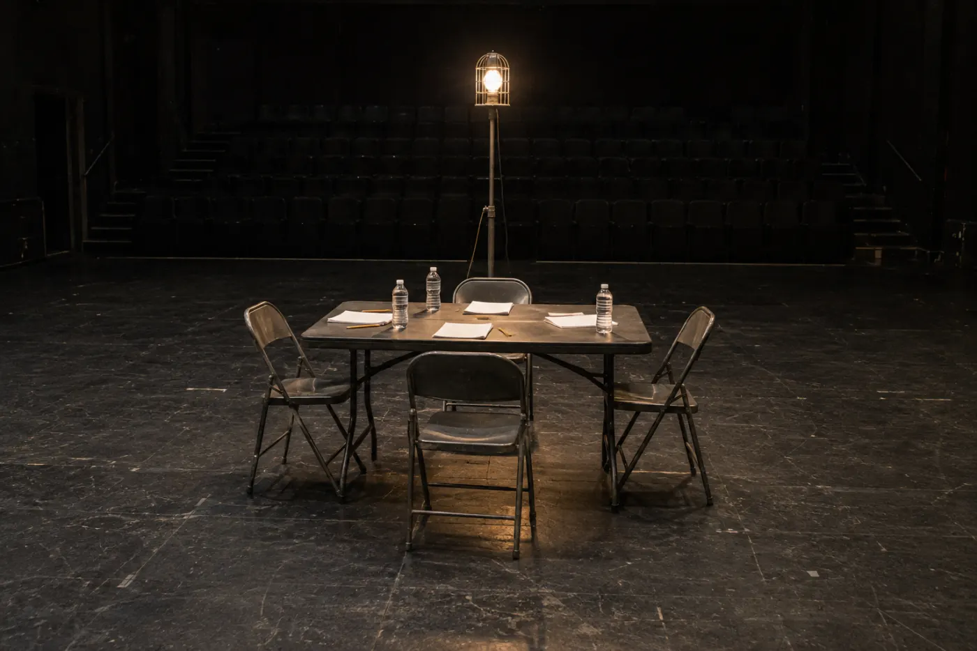 An empty stage with a folding table, scripts, water bottles and four chairs under a single ghost light